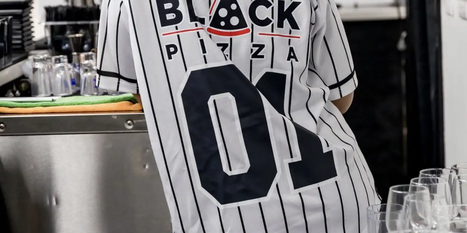 Guest wearing a baseball uniform with sponsorship from Black Pizza on the back. 